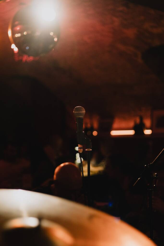 Gros plan sur un microphone sur pied sur une scène faiblement éclairée, avec un reflet de boule disco, évoquant une ambiance de concert live au Caveau.